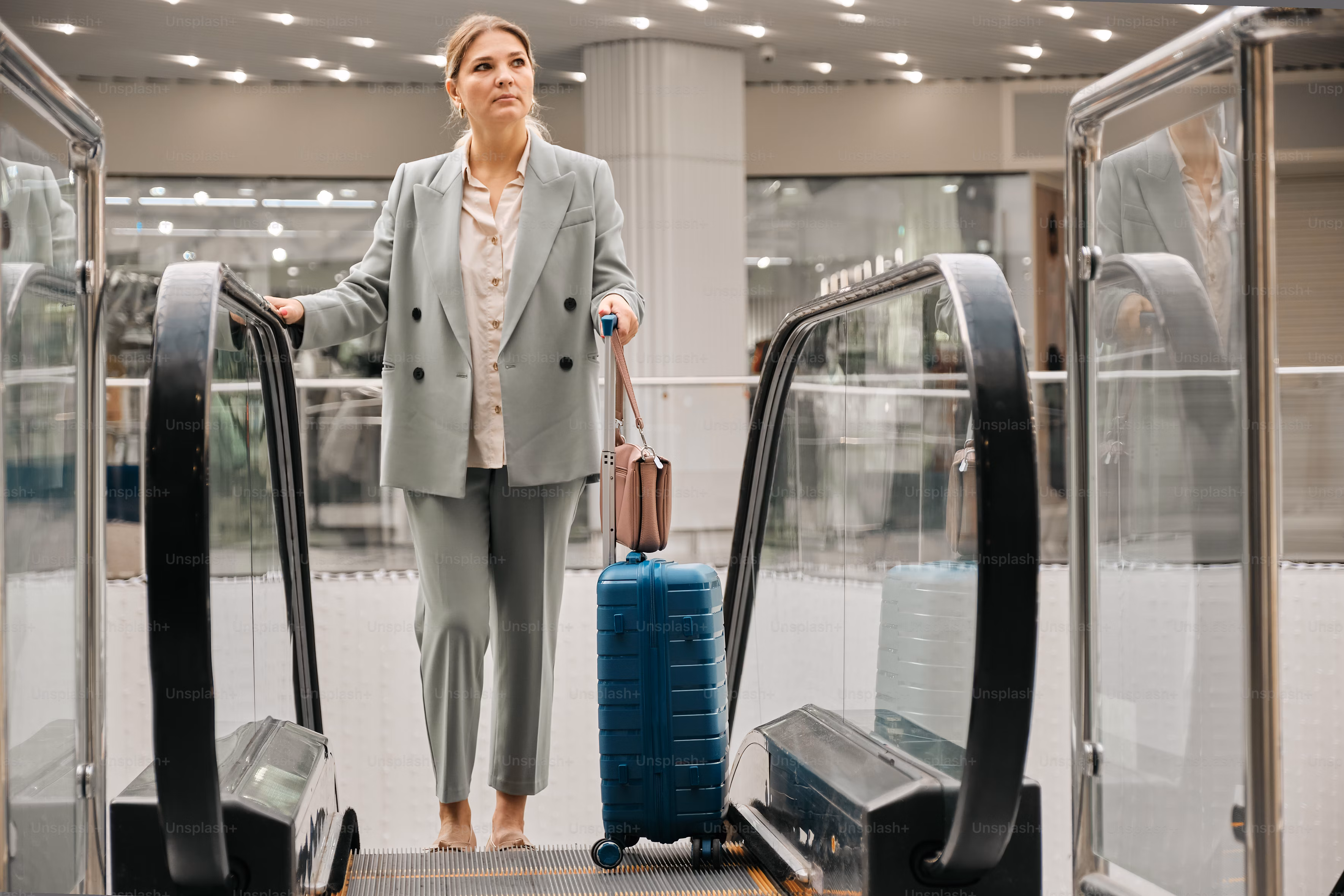 A woman in a suit stands on an escalator with a blue rolling suitcase and a handbag, embodying the dynamic lifestyle of jobs with travel opportunities in a modern indoor setting.