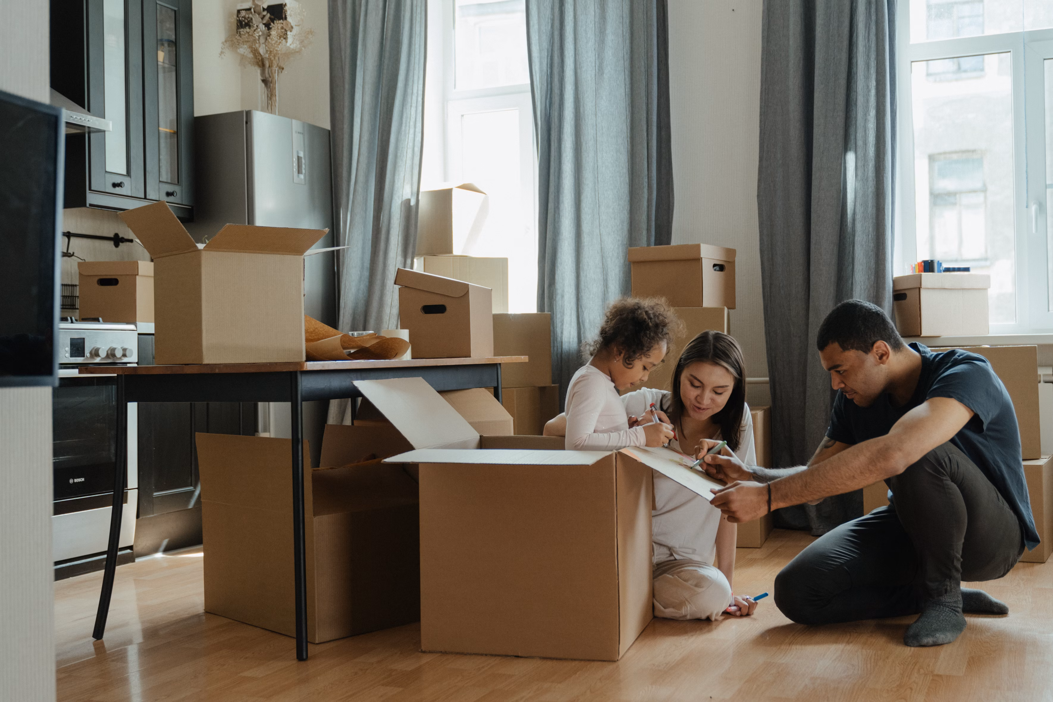 A family unpacks moving boxes in a living room, with a child sitting inside a box while two busy mums read buy a house tips from a document nearby.